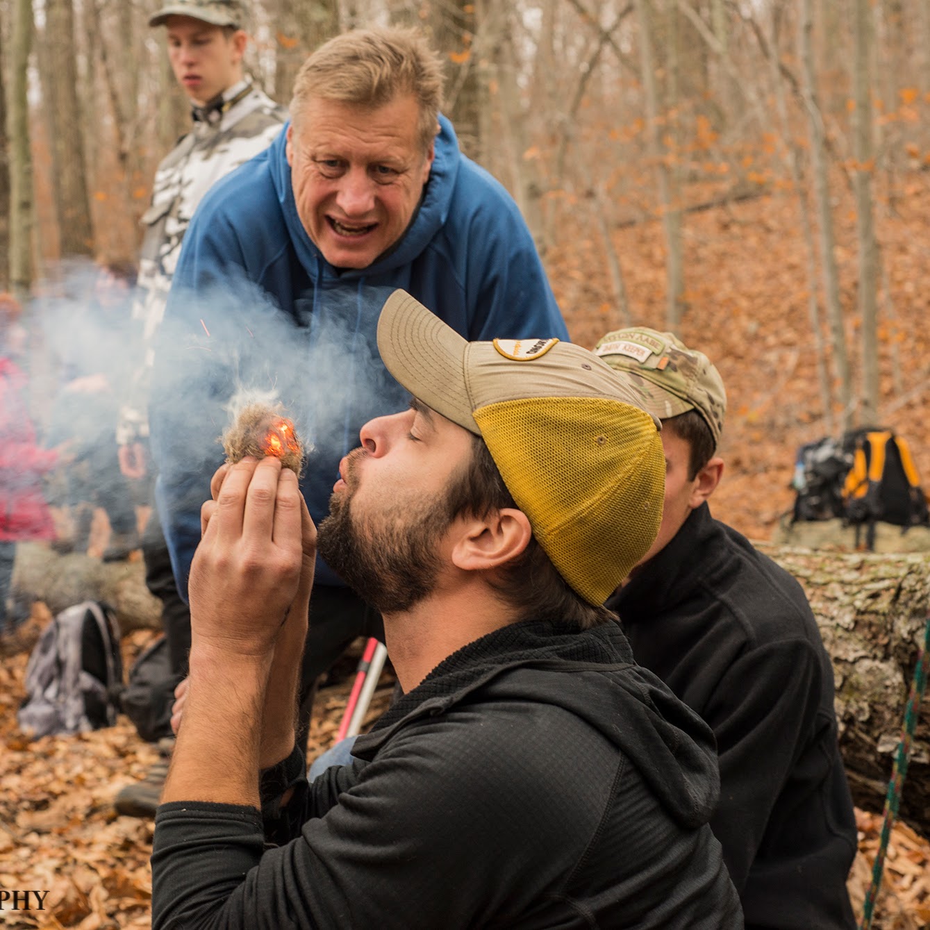 Man blowing into a fire starter in a forest setting with other people around.