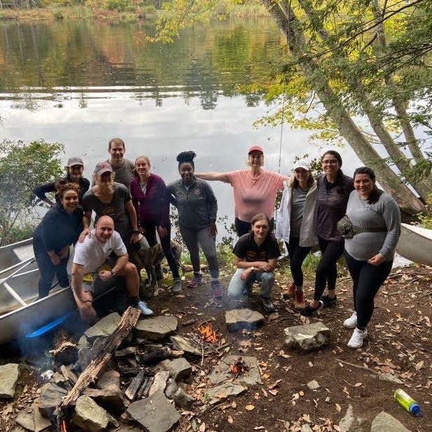 a group poses around a small fire with a mountain lake in the background