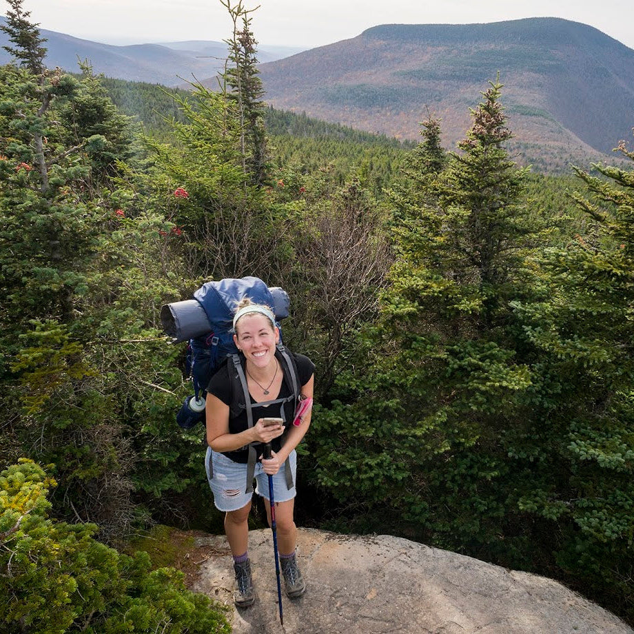 Person hiking with a backpack on a trail surrounded by trees and mountains