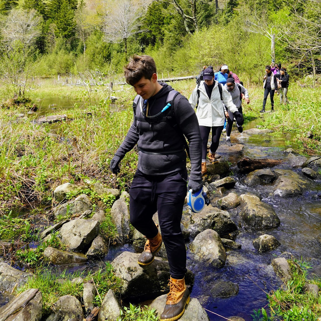 Group of hikers crossing a stream in a forested area with mountains in the background