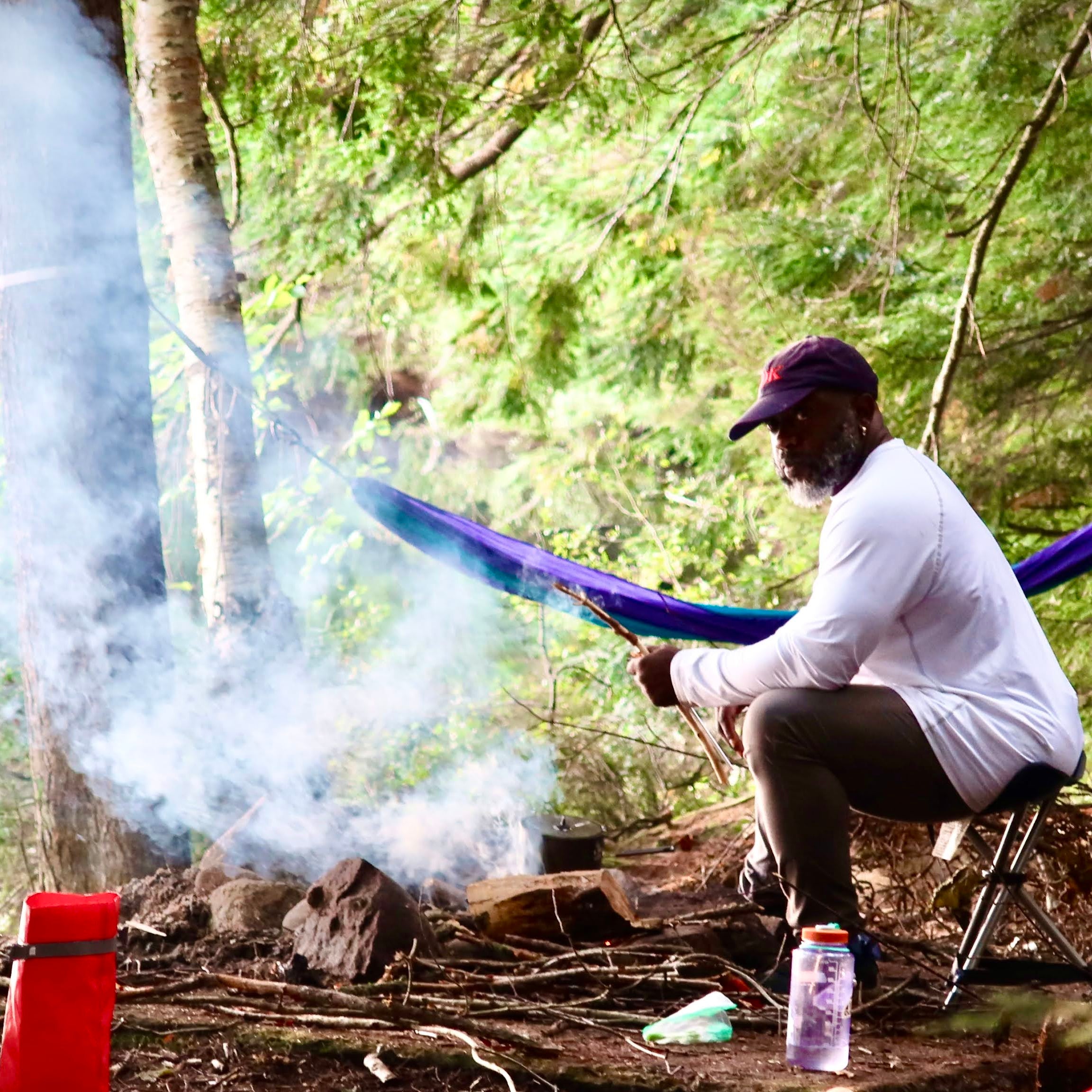 Person sitting in a hammock in a forest with camping gear and smoke