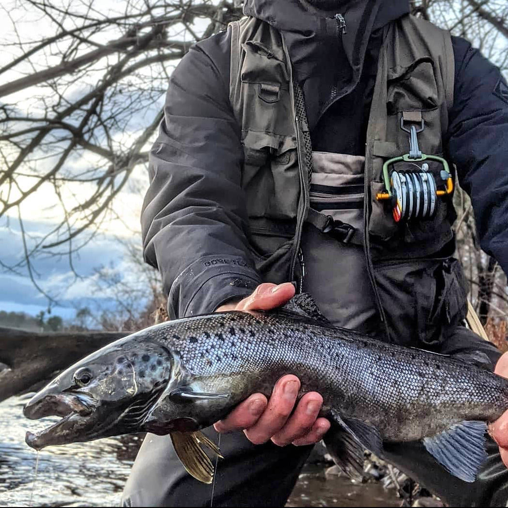 Man holding a large fish by a river with bare trees in the background