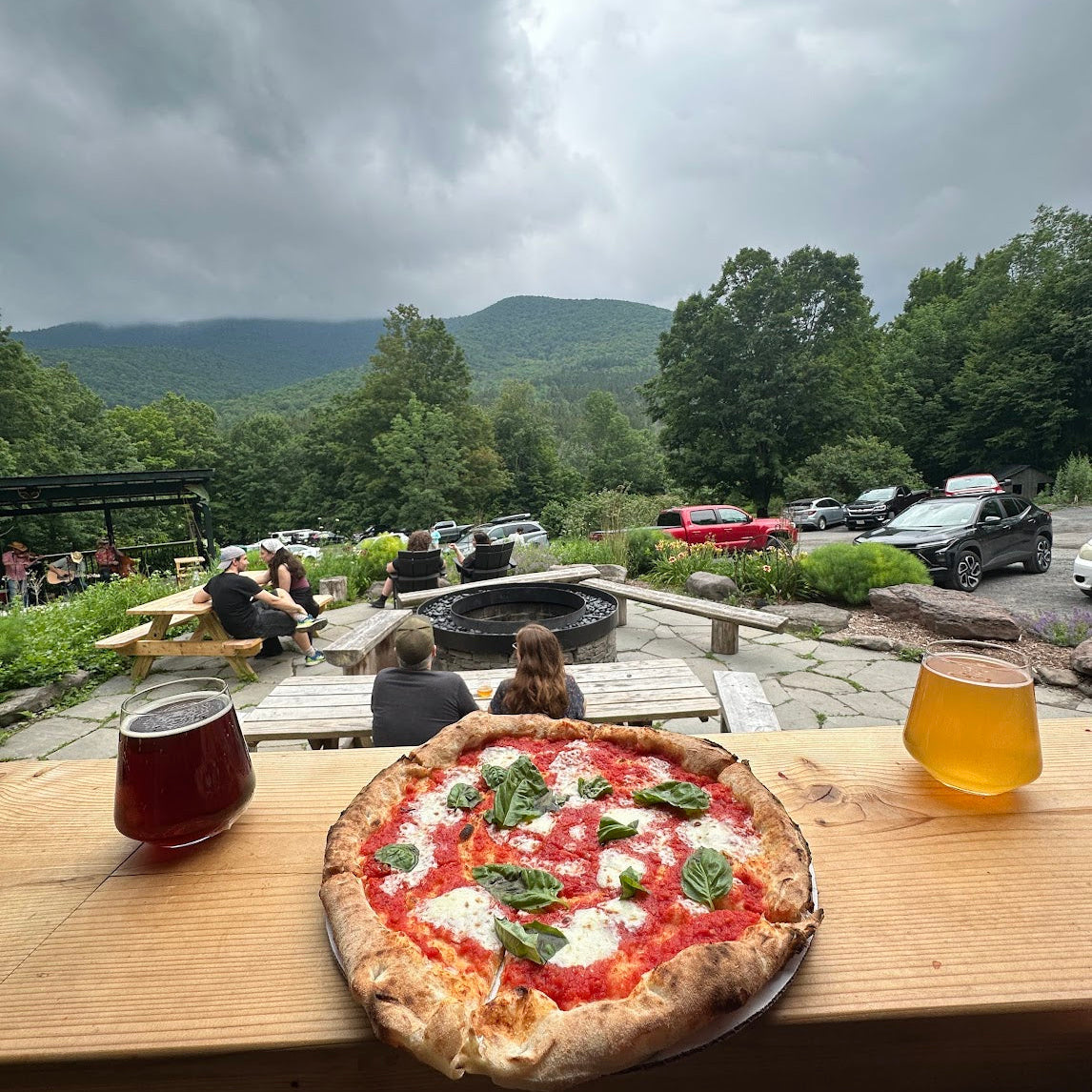 Pizza on a wooden table with drinks, surrounded by a scenic outdoor setting with mountains and trees.
