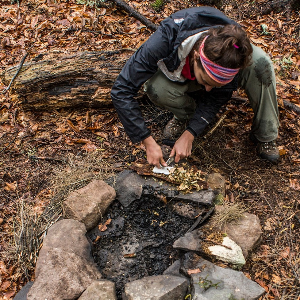 a woman strikes sparks with magensium into a pile of birch bark in front of a fire pit
